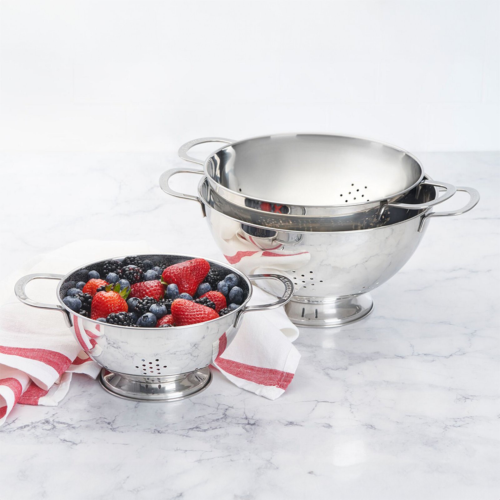 1 photo of 3 stainless steel Le Creuset strainers, 1st filled with strawberries and blueberries, 2nd nested inside of 3rd and empty.